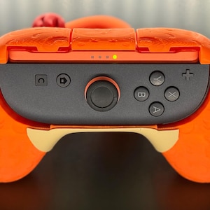 May include: An orange and black video game controller with a unique design. The controller features a central black panel with buttons and a joystick, surrounded by an orange shell. The design includes a textured pattern and a red accent.
