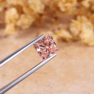 May include: A princess-cut gemstone with a light peach-pink hue held by silver-toned metal tweezers. The stone features a faceted surface that reflects light, creating a brilliant appearance. The watermark crystalcavejewels is visible.