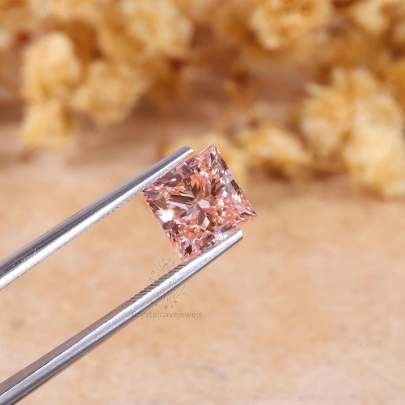 May include: A princess-cut gemstone with a light peach-pink hue held by silver-toned metal tweezers. The stone features a faceted surface that reflects light, creating a brilliant appearance. The watermark crystalcavejewels is visible.