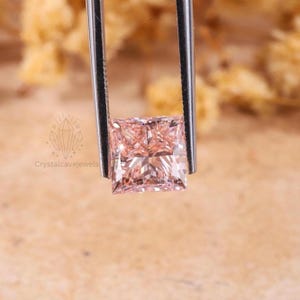 May include: A square-cut, light pink gemstone held by silver-toned metal tweezers. The stone features a faceted surface that reflects light, creating a brilliant appearance. A small logo with the text Crystalcavejewels is visible on the left.