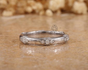 May include: A silver ring with multiple small, clear gemstones. The ring has a rounded band with a series of small gemstones set around the band. The ring also has three larger gemstones set in the center. The ring is on a brown surface.