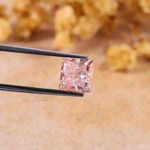 May include: A square-cut peach-colored gemstone held by a pair of metal tweezers. The stone features a faceted surface that reflects light, creating a brilliant appearance. The text Crystalcavejewels is visible in the lower left corner.