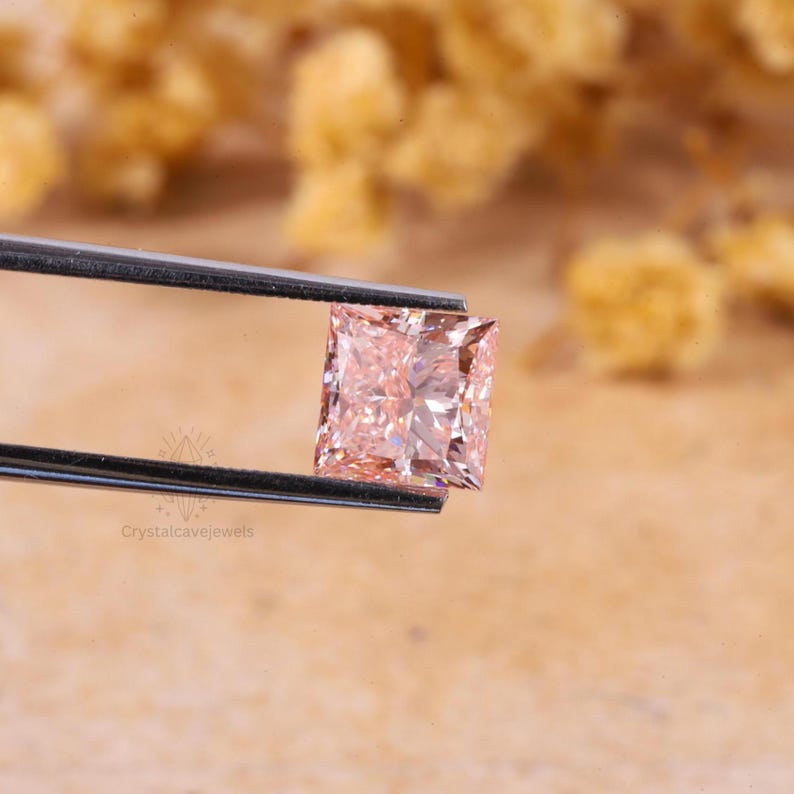 May include: A square-cut peach-colored gemstone held by a pair of metal tweezers. The stone features a faceted surface that reflects light, creating a brilliant appearance. The text Crystalcavejewels is visible in the lower left corner.