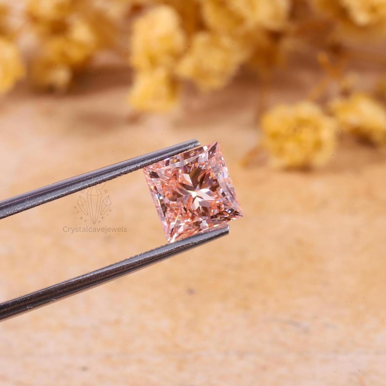 May include: A princess-cut gemstone with a light peach-pink hue held by metal tweezers. The stone features a faceted surface that reflects light, creating a brilliant appearance. The text Crystalcavejewels is visible near the metal tool.