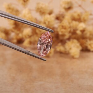 May include: A pink marquise-shaped gemstone held by tweezers. The gemstone is faceted and has a brilliant shine.