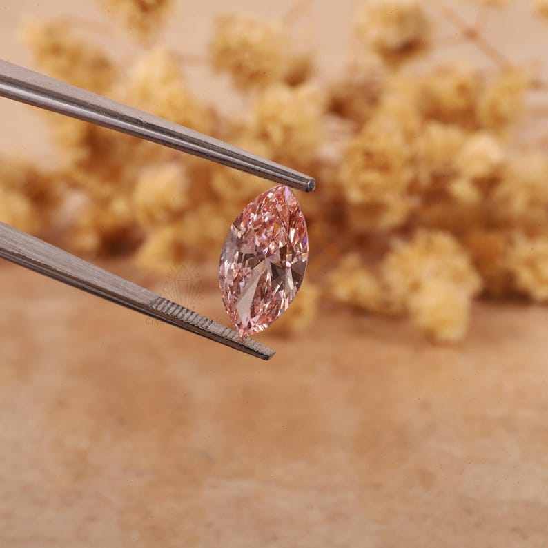 May include: A pink marquise-shaped gemstone held by tweezers. The gemstone is faceted and has a brilliant shine.