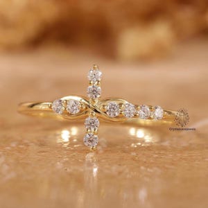 Puede incluir: Un anillo de oro con un diseño central en forma de cruz adornado con pequeñas piedras transparentes de corte redondo. La banda es delgada y pulida, con el diseño de la cruz extendiéndose horizontal y verticalmente por el centro con brillo.