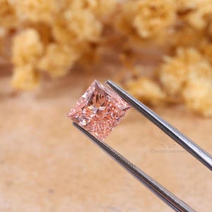 May include: A square-cut, light peach-colored gemstone held by silver-toned metal tweezers. The stone features a faceted surface that reflects light, creating a sparkling appearance. The text Crystalcavejewel is visible on the metal tweezers.