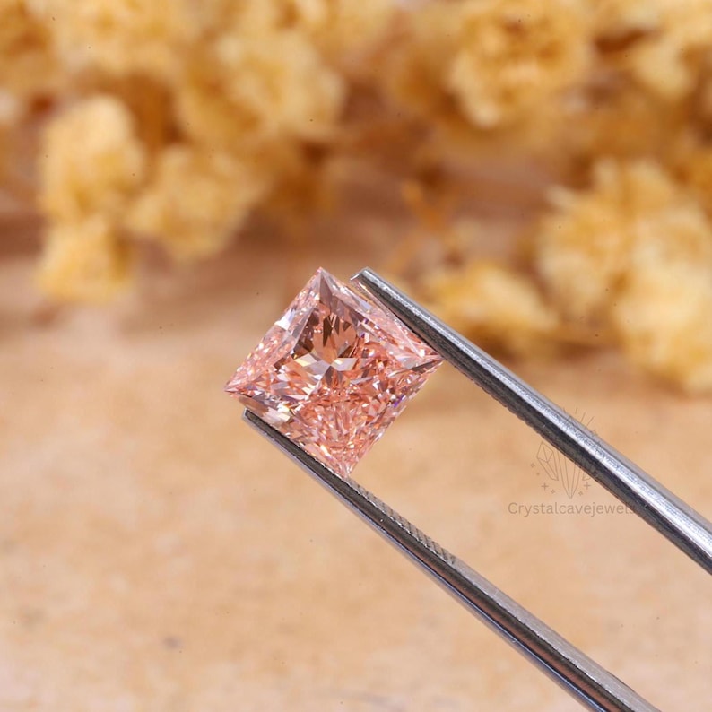 May include: A square-cut, light peach-colored gemstone held by silver-toned metal tweezers. The stone features a faceted surface that reflects light, creating a sparkling appearance. The text Crystalcavejewel is visible on the metal tweezers.