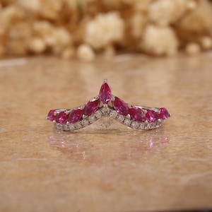 May include: A silver-toned ring with a V-shaped design. The ring features a row of marquise-cut, vibrant pink gemstones and smaller, clear stones. The ring is set on a neutral background, with the text "crystalcavejewels" visible.