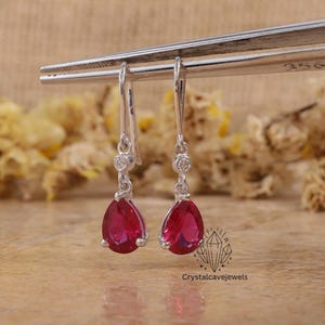 May include: Silver drop earrings with vibrant, pear-cut red gemstones. Each earring has a small, round accent stone above the red gem. The earrings are displayed by a silver tool against a neutral backdrop.