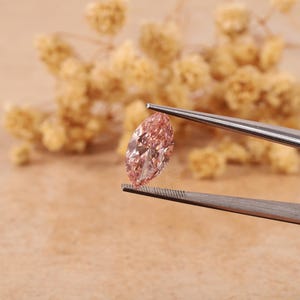 May include: A pink marquise-shaped diamond gemstone held in tweezers. The diamond is clear and has a slight pink hue.
