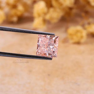 May include: A square-cut, light peach-colored gemstone held by a pair of metal tweezers. The stone features a faceted surface that reflects light, creating a brilliant appearance. The text Crystalcavejewels is visible on the lower left side.
