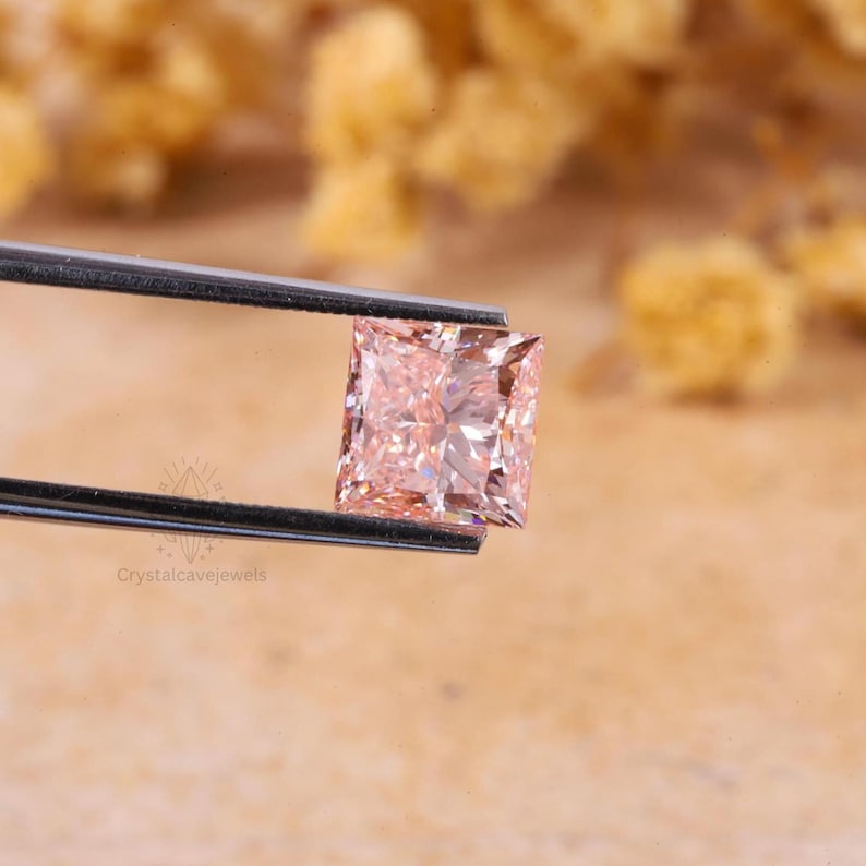 May include: A square-cut, light peach-colored gemstone held by a pair of metal tweezers. The stone features a faceted surface that reflects light, creating a brilliant appearance. The text Crystalcavejewels is visible on the lower left side.