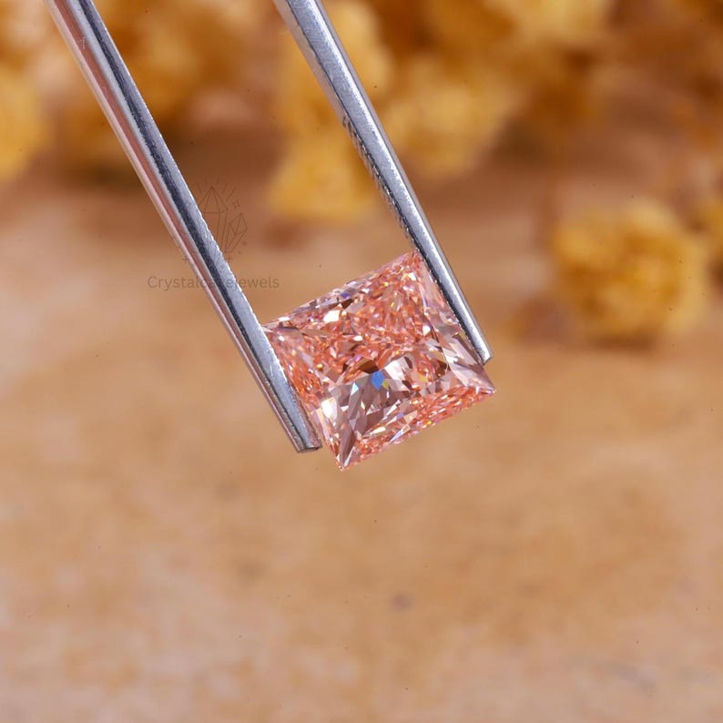 May include: A square-cut, light peach-colored gemstone held by silver-toned metal tweezers. The stone features a faceted surface that reflects light, creating a sparkling appearance. The text Crystal Jewels is visible on the stone surface.
