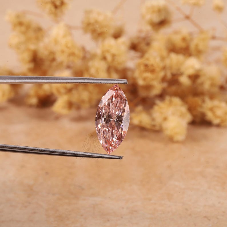 May include: A light pink marquise-shaped gemstone held by tweezers. The gemstone has a brilliant cut and facets that reflect light.