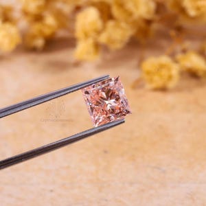 May include: A princess-cut gemstone with a light peach-pink hue held by metal tweezers. The stone features a faceted surface that reflects light, creating a brilliant sparkle. The logo Crystalcavejewels is visible on the metal arm of the tweezers.