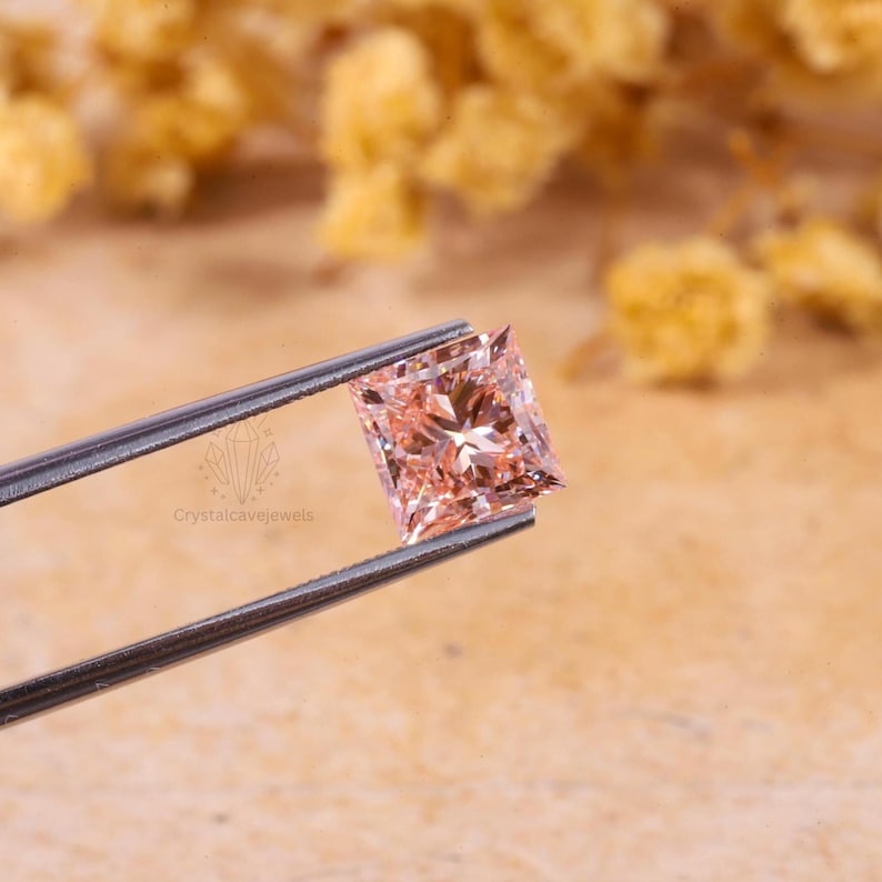 May include: A princess-cut gemstone with a light peach-pink hue held by metal tweezers. The stone features a faceted surface that reflects light, creating a brilliant sparkle. The logo Crystalcavejewels is visible on the metal arm of the tweezers.