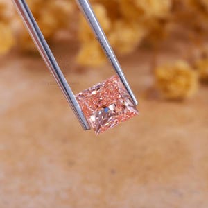 May include: A square-cut peach-colored gemstone held by silver-toned metal tweezers. The stone features a faceted surface that reflects light, creating a brilliant appearance. The text CrystalJewels is visible on the metal of the tweezers.