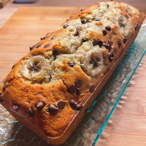 May include: A loaf of banana bread with chocolate chips on top. The bread is golden brown and has a slightly crispy crust. The bread is sitting on a clear glass plate.