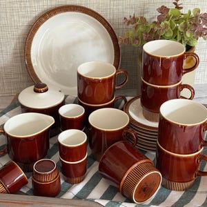 May include: A collection of brown and cream ceramic tableware including mugs, a lidded sugar bowl, a creamer, and a plate. The mugs feature a glossy brown glaze on the upper portion and a ribbed, matte brown texture on the base. Each piece is marked Poole.