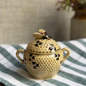 May include: A ceramic sugar bowl with a honeycomb texture in shades of tan and yellow, featuring black clover accents and two side handles. The lid is topped with a sculpted bee figure, and the entire piece has a glossy finish throughout.