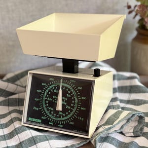 May include: A cream-coloured EKS kitchen scale featuring a large, removable top tray and a black display panel. The dial shows dual measurements in pounds and kilograms with green text and a white needle. The scale has a compact, boxy design.