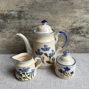 May include: A cream ceramic tea set with blue floral patterns. The set comprises a teapot, a creamer jug, and a lidded sugar bowl. The teapot has a long spout and a blue handle. The floral design features blue flowers with green stems.