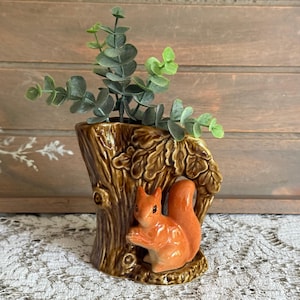 May include: A brown ceramic vase shaped like a tree trunk with a sculpted orange squirrel attached to the front. The vase features textured bark details and oak leaves. A sprig of green artificial eucalyptus leaves is placed inside the vase.
