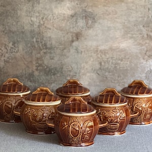 May include: A collection of six brown ceramic lidded pots with handles. Each pot features a detailed relief design of a tree trunk with a squirrel and other forest elements. The lids have a ridged texture.