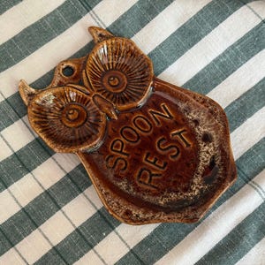 May include: A brown glazed ceramic spoon rest shaped like an owl with large circular eyes and pointed ears. The words SPOON REST are embossed in the centre of the body. The finish features a glossy, mottled brown texture throughout the piece.