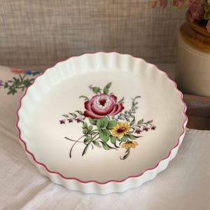 May include: A round white ceramic tart dish with a fluted edge and a thin pink rim. The centre of the dish features a detailed floral illustration with a large dark pink rose, smaller yellow flowers, and green leaves on a white background.
