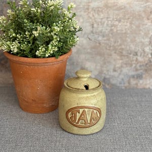 May include: A ceramic jar with a lid, decorated with brown speckles and the word "JAM" in brown lettering. The jar is sitting on a grey textured surface.