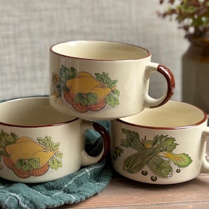 May include: Three cream-coloured ceramic soup mugs with brown handles and rims are stacked. Each mug features a different illustration: a roasted chicken, and celery. The mugs are on a wooden tray.