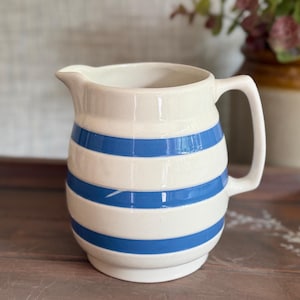 May include: A cream-coloured ceramic jug featuring three horizontal blue stripes around its rounded body. The jug has a wide pouring spout, a sturdy handle on the right side, and a smooth, glossy finish throughout its exterior surface.
