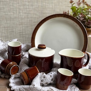 May include: A vintage brown and white ceramic tableware set. The set includes a large plate, sugar bowl, creamer jug, and several small cups. The pieces have a brown band design and are arranged on a white lace cloth.