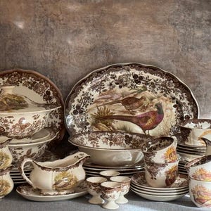 May include: A collection of vintage brown and white ceramic tableware. The set includes serving dishes, bowls, cups, saucers, and a gravy boat. The pieces feature a pheasant and floral design, with a scalloped edge.