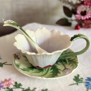 May include: A ceramic teacup and saucer set shaped like a white flower with green leaves and a red base. The teacup features a scalloped rim and a green handle. A matching spoon with a small white flower at the end rests inside the teacup.