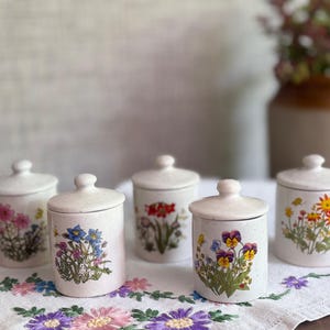 May include: Five small, off-white ceramic jars with lids, each decorated with a different floral design in various colours. The jars are arranged on a white embroidered cloth with a floral pattern. A brown ceramic vase with flowers is in the background.