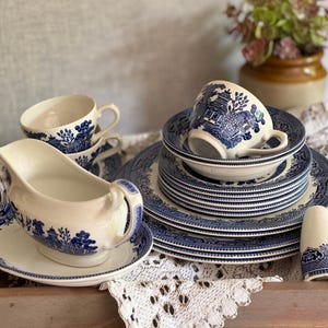 May include: A vintage blue and white ceramic tableware set, featuring plates, cups, a gravy boat, and salt and pepper shakers. The Willow pattern design decorates the items. The set is arranged on a wooden tray with a lace doily.