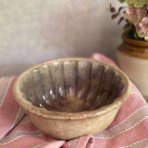 May include: A round, fluted ceramic bowl with a weathered, mottled beige and brown finish. The interior features vertical ridges radiating from the centre, while the exterior has a smooth, rounded rim and a textured, aged appearance.