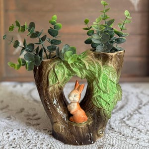 May include: A ceramic double planter shaped like a tree trunk with a brown bark texture and green leaves. A small orange rabbit figurine sits in the centre opening between the two trunks. Two green artificial eucalyptus stems are placed inside.