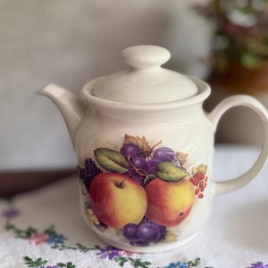 May include: Cream ceramic teapot with a lid and handle. The teapot is decorated with a colourful fruit design featuring apples, grapes, and leaves. The teapot is sitting on a white embroidered cloth.