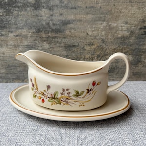 May include: Cream-coloured gravy boat with a matching saucer. The boat has a handle and a spout, and is decorated with a floral and berry design in green, brown, and red. The rim of both pieces is trimmed with a thin brown line. A classic tableware item.