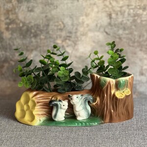 May include: A ceramic planter shaped like a log and tree stump, with two squirrel figurines. The planter is brown, yellow, and green, with green plants inside. The squirrels are white and grey.