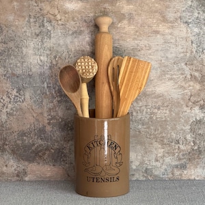 May include: A brown ceramic utensil holder with the words "Kitchen Utensils" printed on the front. The holder contains a wooden rolling pin, a wooden spoon, a wooden spatula, and a wooden whisk.