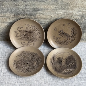 May include: A set of four small, round plates with brown animal illustrations. Each plate features a different animal: a cat, a mouse, two rabbits, and two birds. The plates have a neutral, earthy tone.