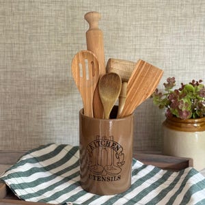 May include: A cylindrical brown ceramic utensil holder with the black text KITCHEN UTENSILS and a line drawing of kitchen tools. It contains a wooden rolling pin, a slotted spoon, a solid spoon, a meat mallet, and a flat wooden spatula.