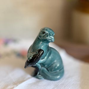 May include: A glossy, teal-coloured ceramic figurine of an otter in a seated position. The otter is holding a smooth, dark, black-coloured stone between its front paws. The sculpture features a reflective, glazed finish with visible contours.