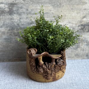 May include: A small, round, tan ceramic planter with a detailed mushroom and foliage design. The planter is filled with vibrant green artificial plants, creating a natural aesthetic. The planter is suitable for small plants and adds a touch of nature to any space.